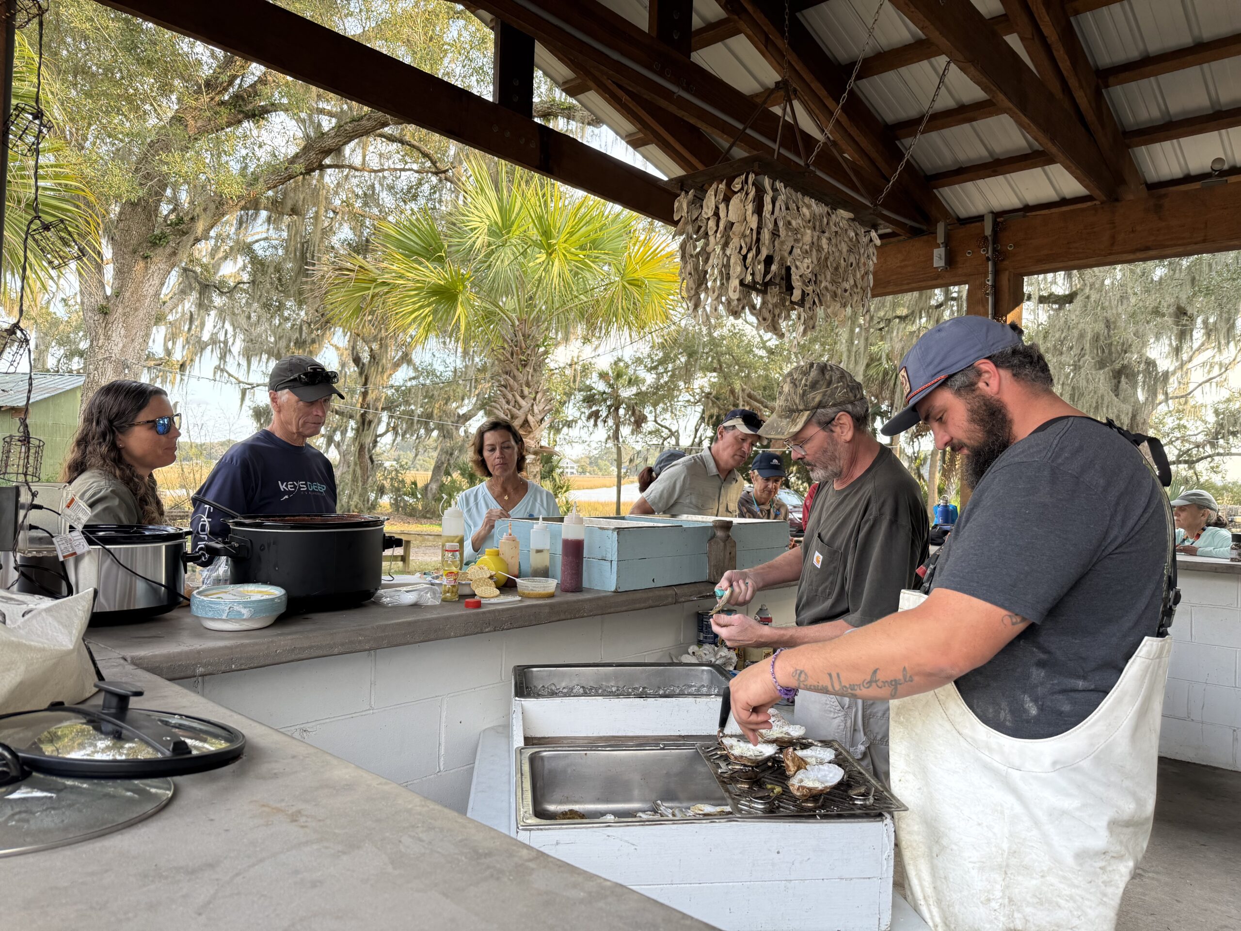 Group of people participating in a seafood cooking event outdoors, with two individuals shucking oysters at a preparation station while others observe and engage in conversation. The setting features a rustic kitchen with palm trees and hanging decorations in the background, highlighting a communal cooking experience.