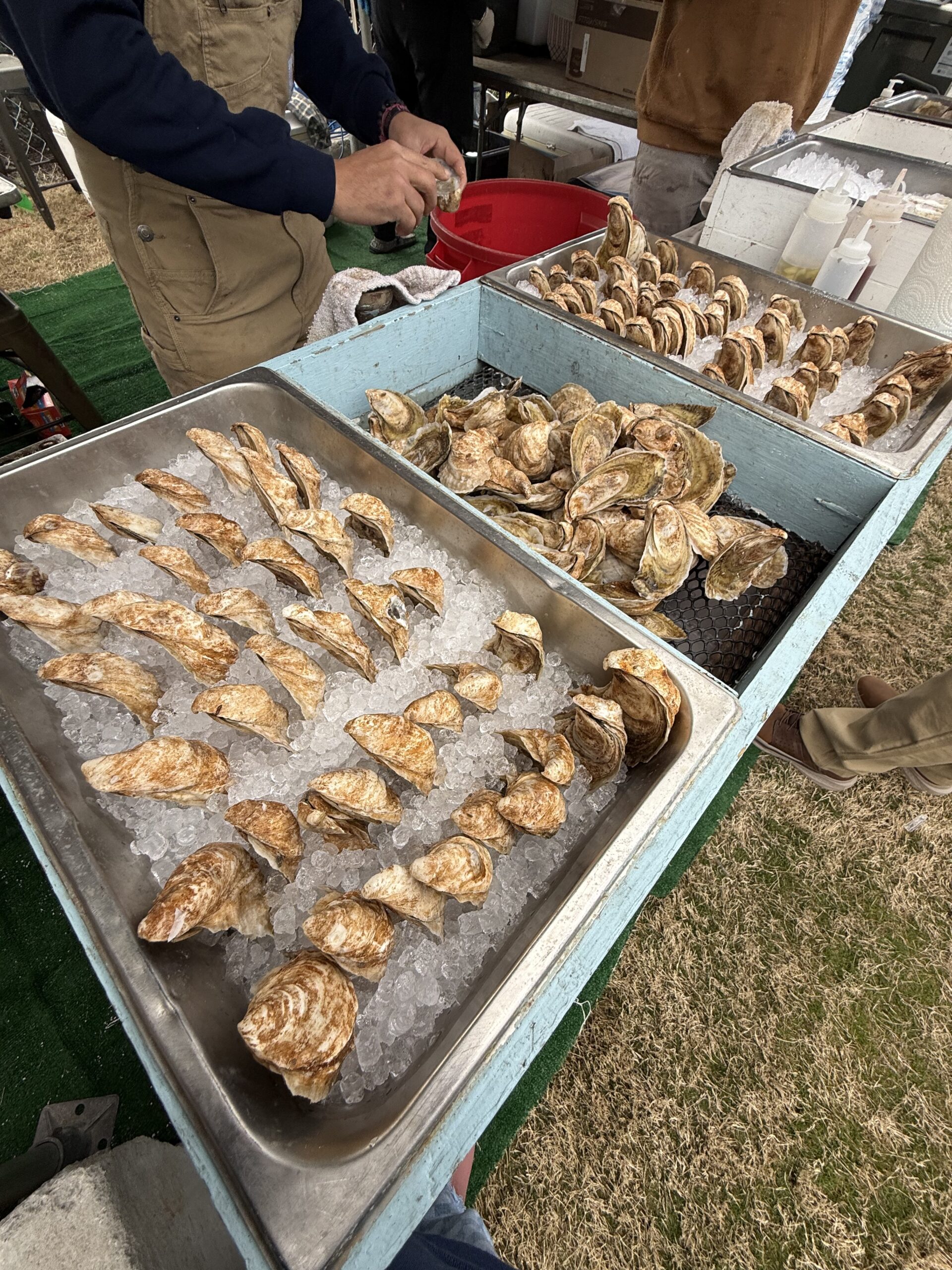 Freshly shucked oysters displayed on ice at a seafood market, with a vendor preparing more oysters in the background.