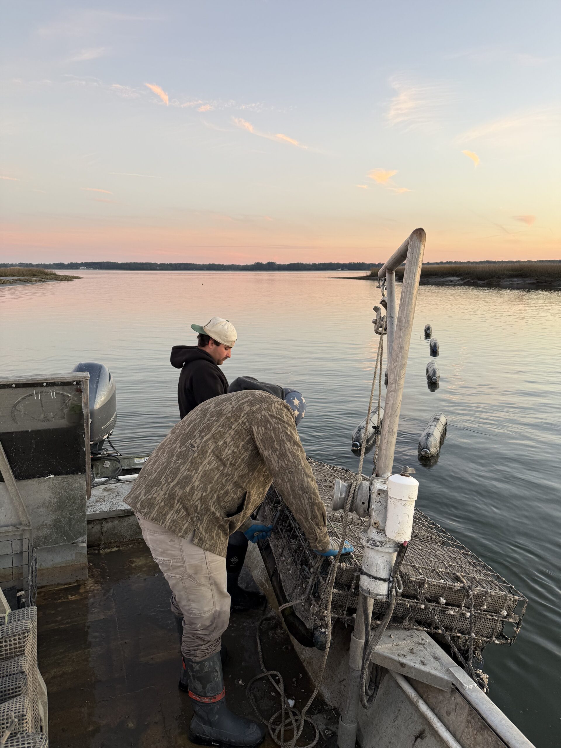 Two fishermen working on a boat during sunset, preparing fishing gear in calm waters. The scene captures a peaceful moment on the water, highlighting the fishing industry.