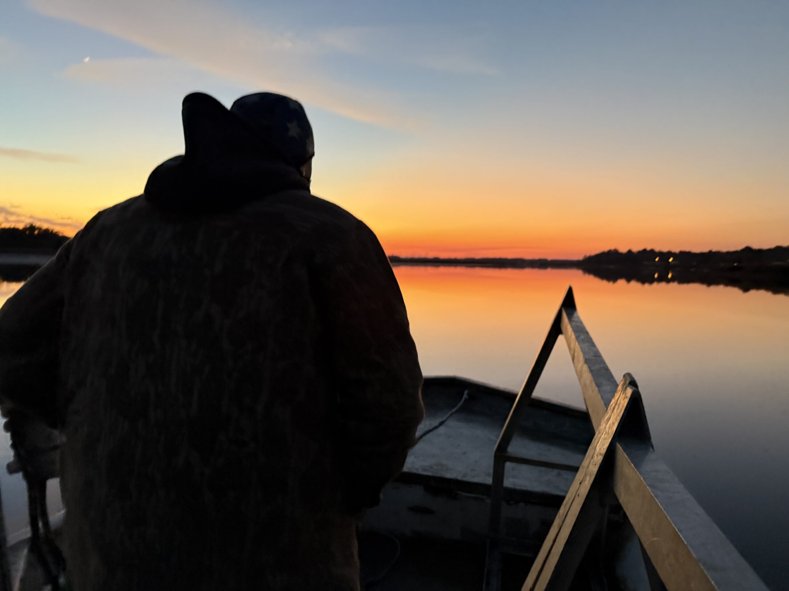 A person in a camouflage jacket stands in a boat at sunrise, overlooking a calm river reflecting vibrant orange and purple hues in the sky, with hints of distant lights along the shoreline.