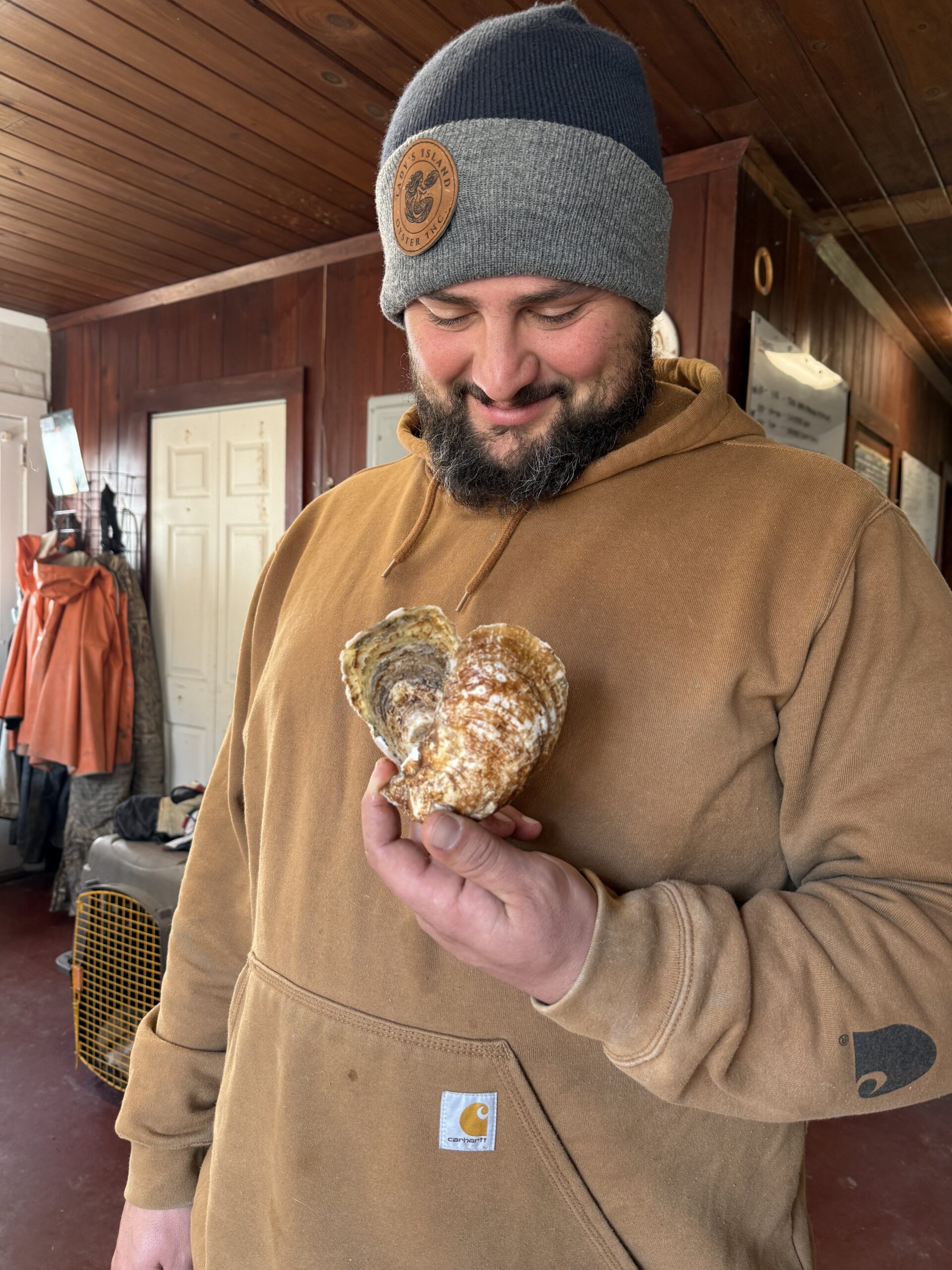 Man holding a large oyster in a rustic setting, wearing a gray and navy beanie and a brown sweatshirt. The background features wooden walls and fishing gear, highlighting a coastal lifestyle.