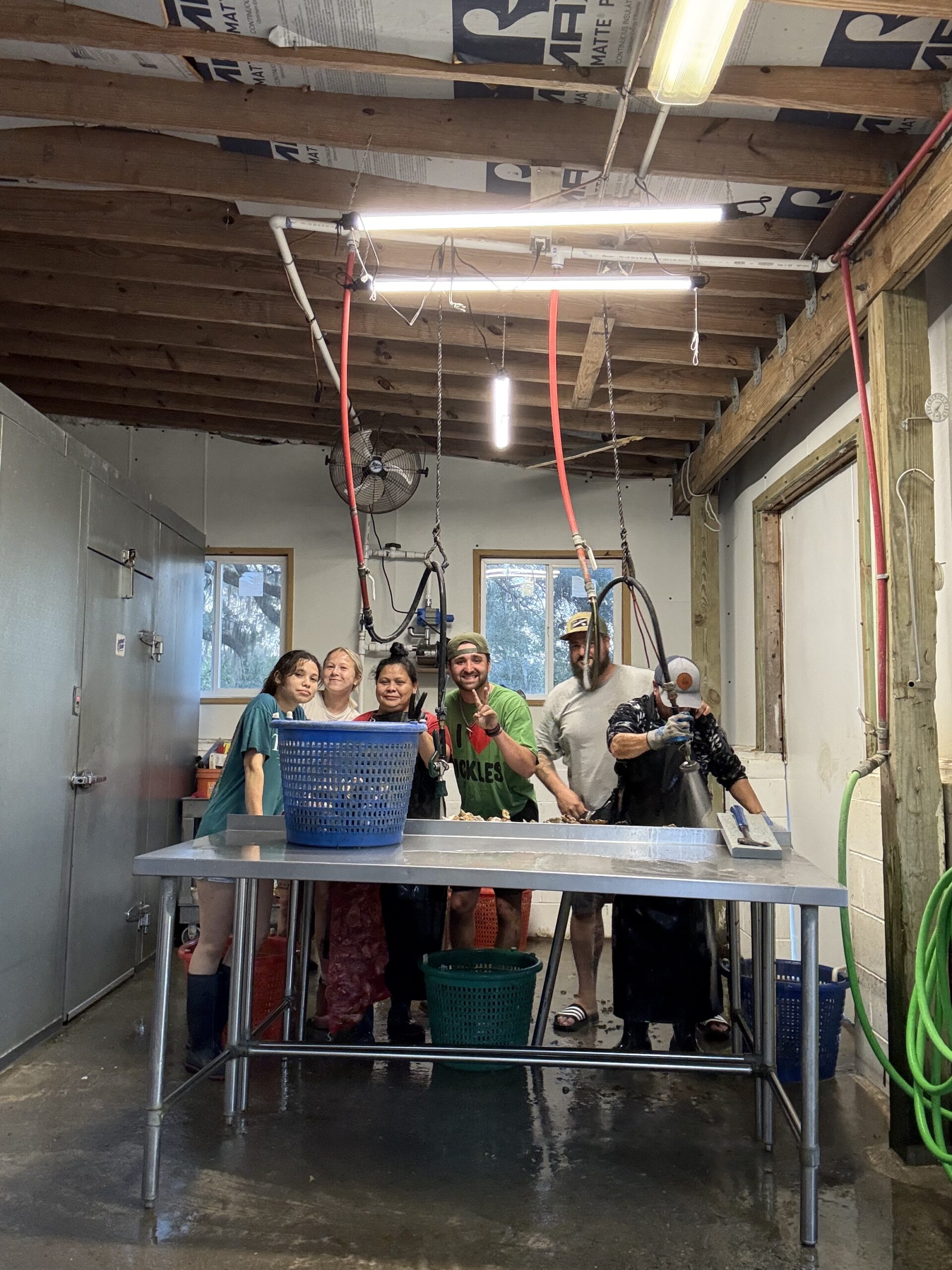 Group of six people in a seafood processing facility, standing around a stainless steel table with blue and green baskets. They are wearing aprons and gloves, smiling, with equipment and water hoses visible in the background, showcasing a vibrant work environment.