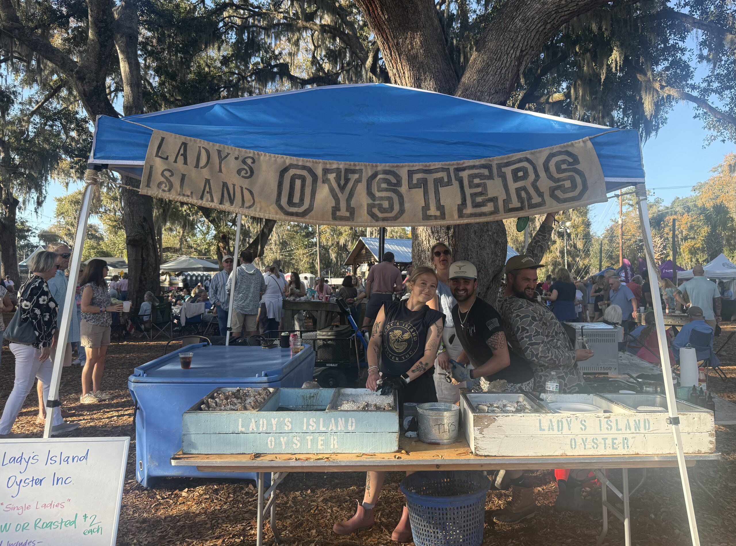 Lady's Island Oysters stand at a local event, featuring vendors serving fresh oysters. The scene includes a bustling crowd, with tables set up under a blue tent and a rustic sign. Three staff members are seen preparing oysters, showcasing the vibrant atmosphere of the festival.